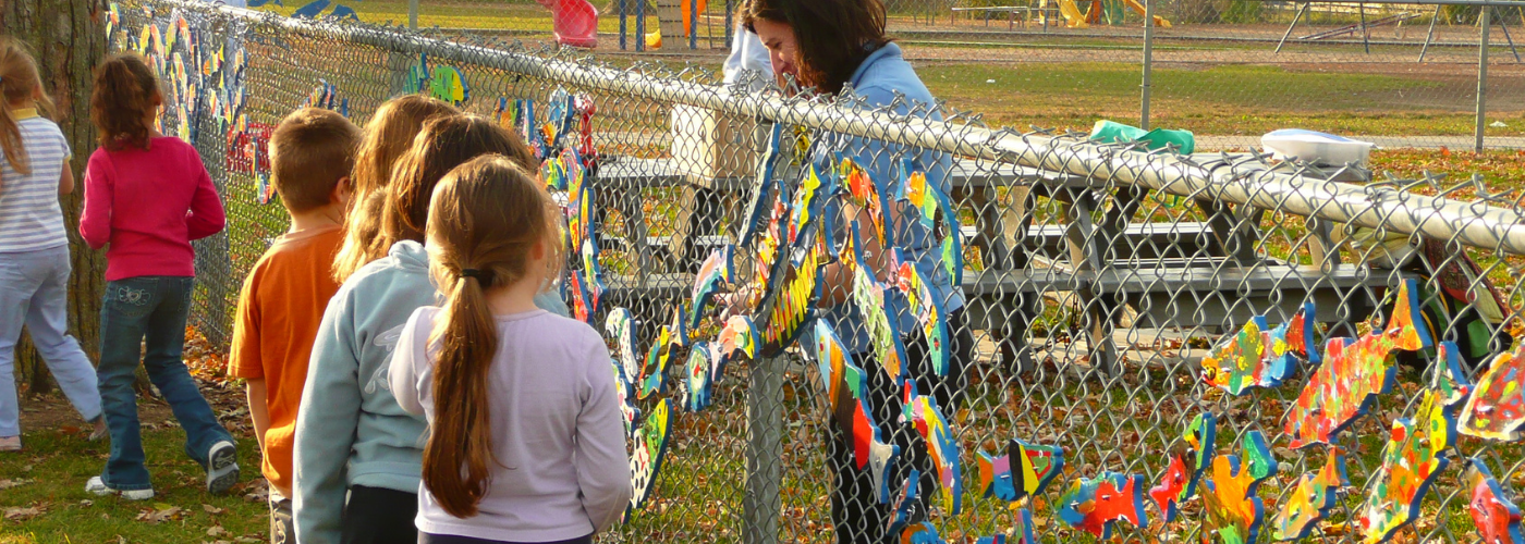 School children walk along a chain link fence with a recently installed stream of dreams mural, painted wooden fish cutouts fixed to the fence in an art mural..